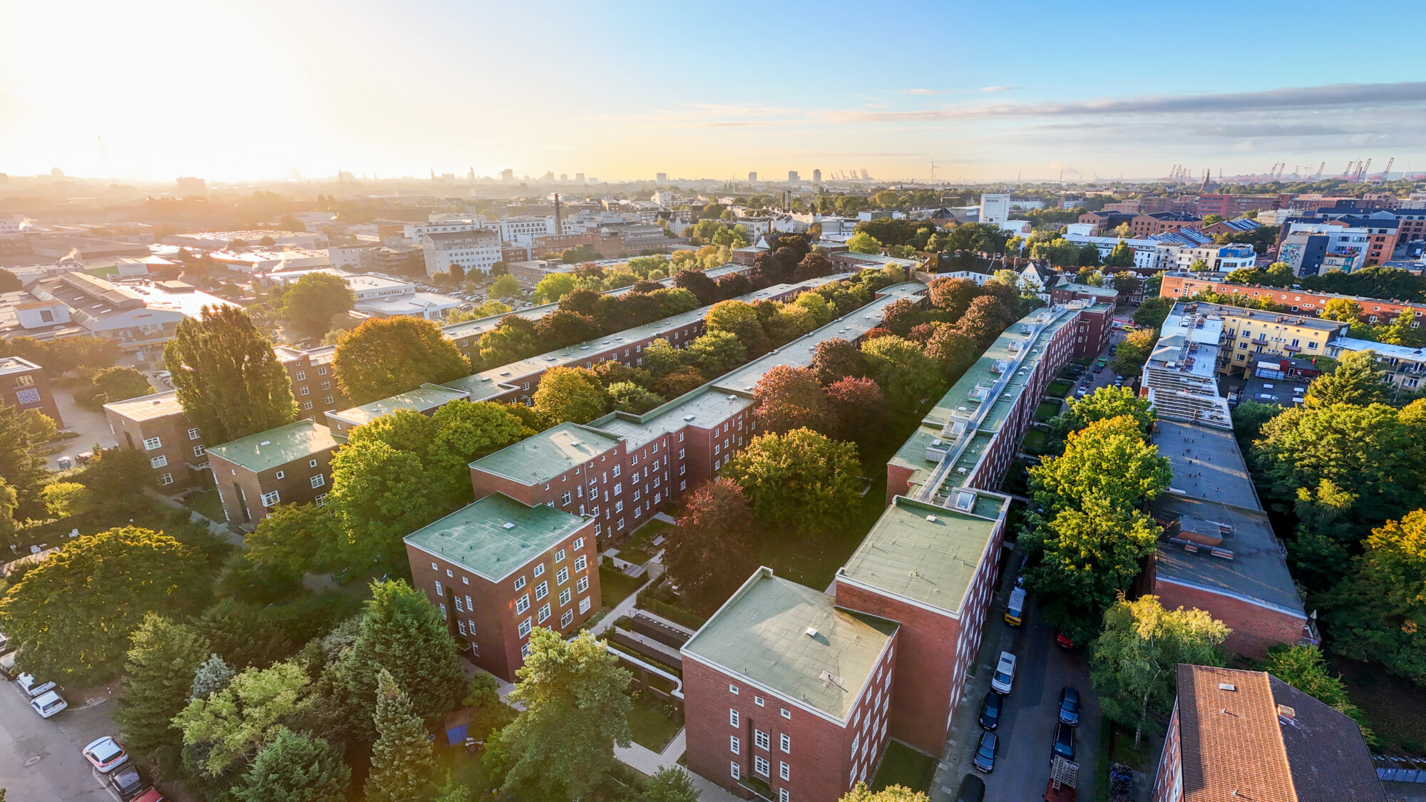 Luftaufnahme einer großen Wohnanlage mit mehreren viergeschossigen Backsteinbauten in U-Form, umgeben von Bäumen und Straßen; im Hintergrund Stadtansicht mit Industrie- und Hafenanlagen.
