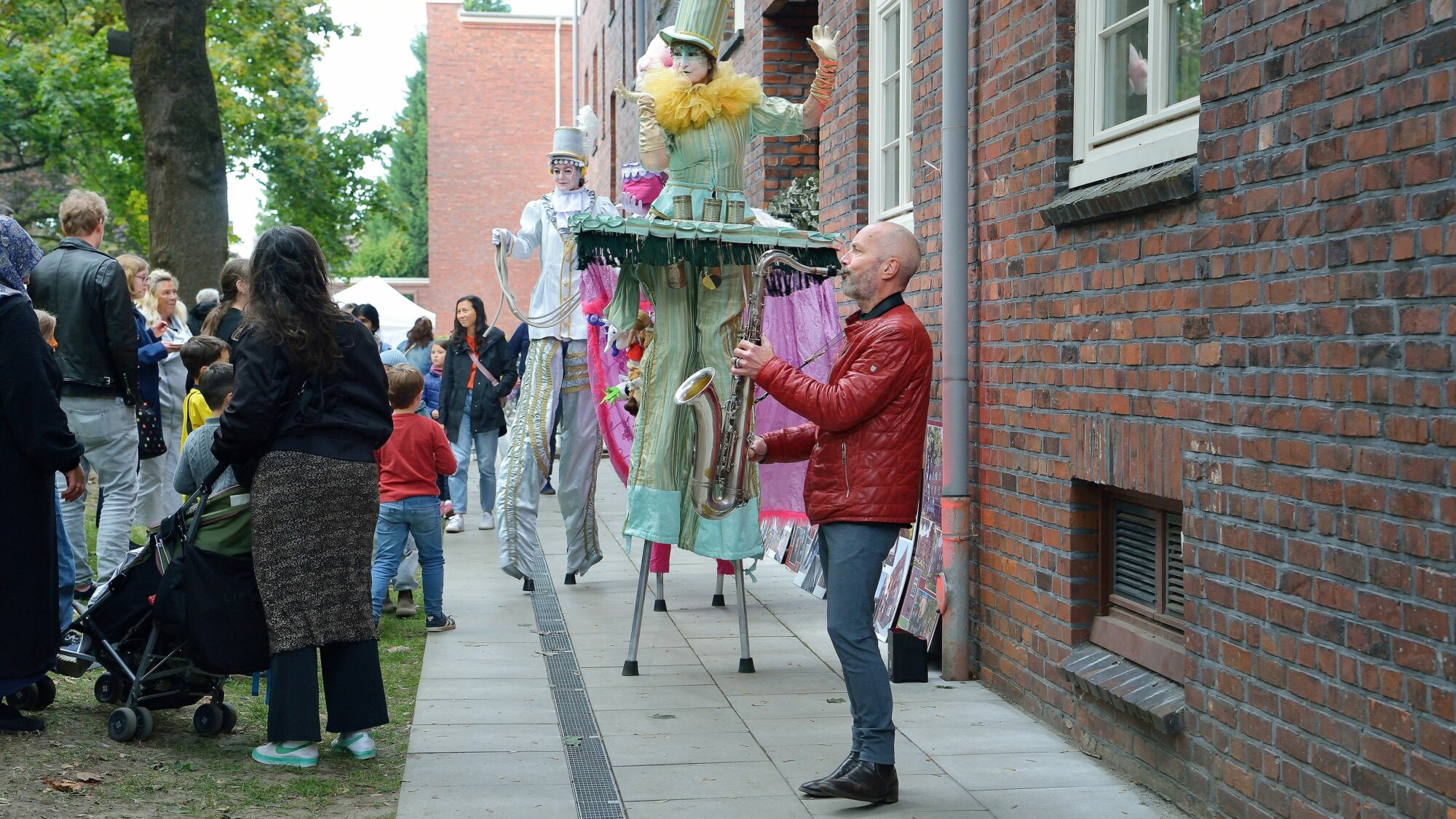 Straßenfest vor einem Backsteingebäude mit verkleideten Künstler:innen auf Stelzen, einem Saxophonspieler und zahlreichen zuschauenden Kindern und Erwachsenen.