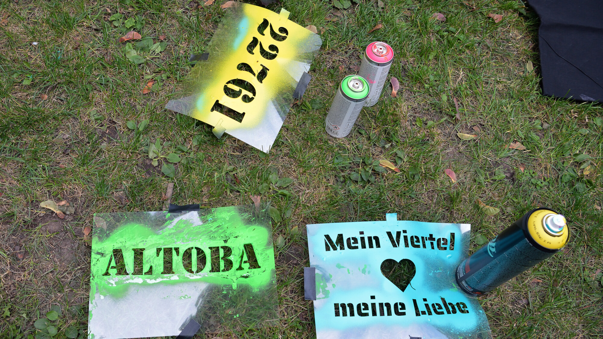 Barrierefrei Bildbeschreiber sagte: Sprühschablonen mit den Aufschriften „ALTOBA“, „1927“ und „Mein Viertel ♥ meine Liebe“ liegen auf einer Wiese, daneben mehrere Farbspraydosen.