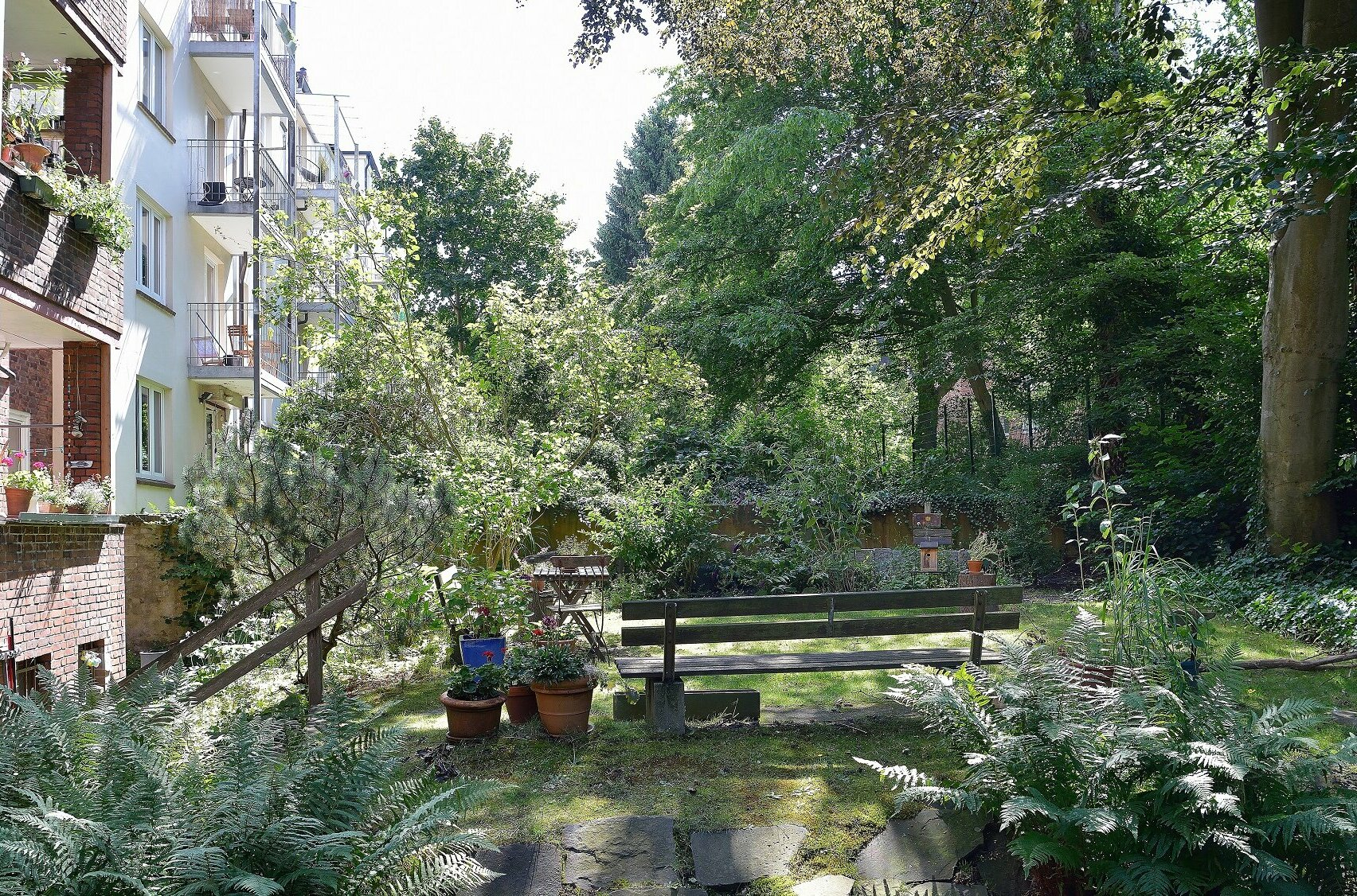 Begrünter gemeinschaftlicher Garten hinter Wohnhäusern mit Sitzbank, Pflanzkübeln und üppiger Vegetation, links Balkone mit Blumenschmuck.