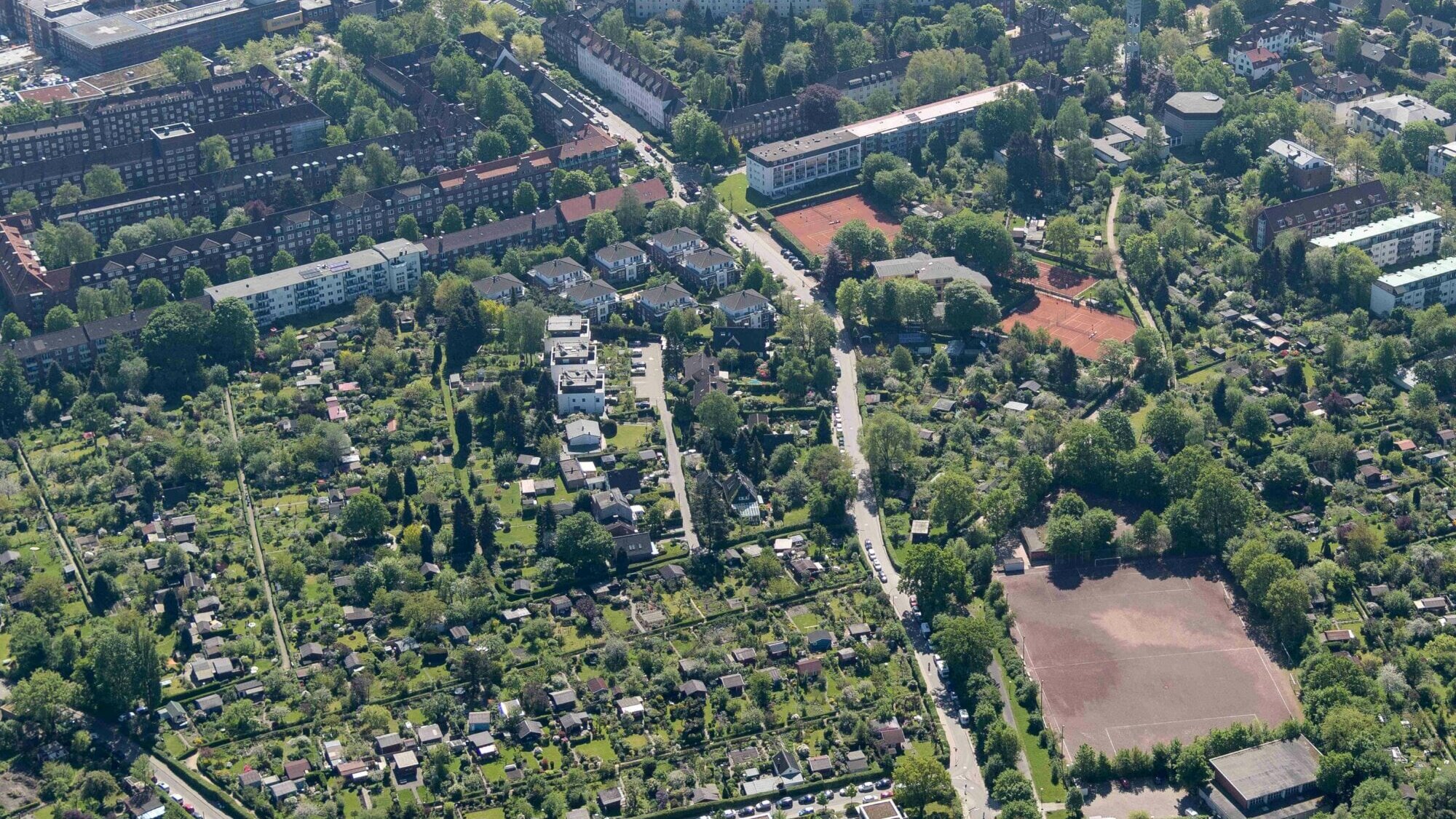 Luftaufnahme eines städtischen Wohngebiets mit dichter Bebauung, Kleingartenanlagen, mehreren Sportplätzen und durchgrünten Straßenzügen inmitten vieler Bäume.