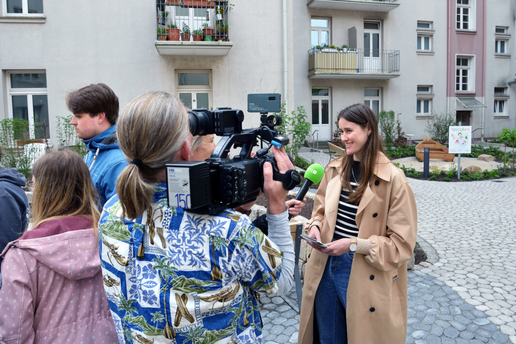 Frau in beigefarbenem Mantel wird im Innenhof eines Wohnhauses von einem Kamerateam interviewt; im Hintergrund ist ein Schild mit der Aufschrift „Spielplatz“ zu sehen.