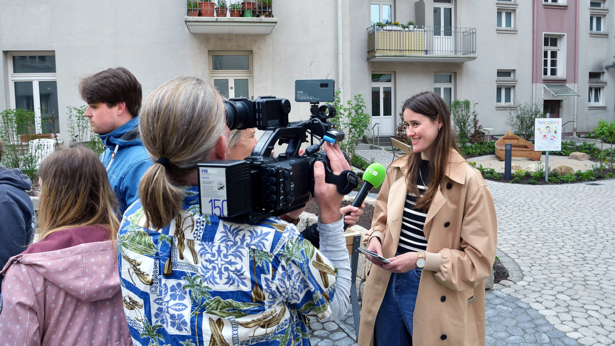 Frau in beigefarbenem Mantel wird im Innenhof eines Wohnhauses von einem Kamerateam interviewt; im Hintergrund ist ein Schild mit der Aufschrift „Spielplatz“ zu sehen.