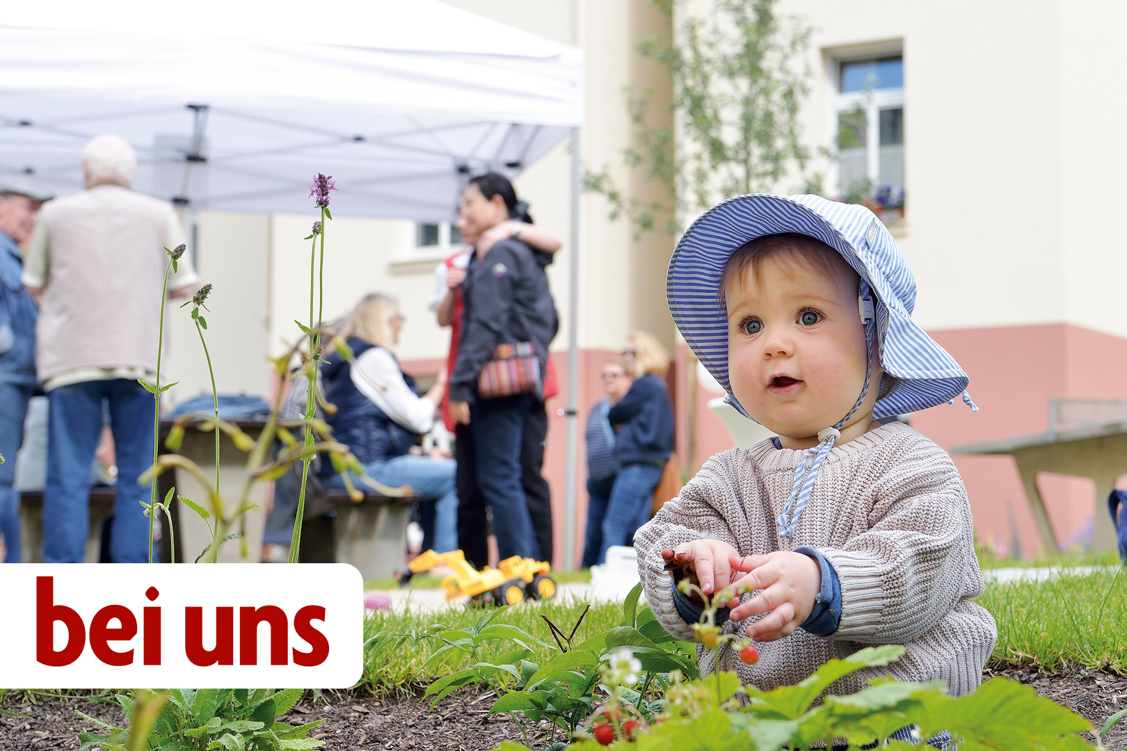 Kleines Kind mit Sonnenhut sitzt auf einer Wiese und pflückt Erdbeeren; im Hintergrund Menschen unter Pavillons bei einem Nachbarschaftstreffen oder Hoffest.