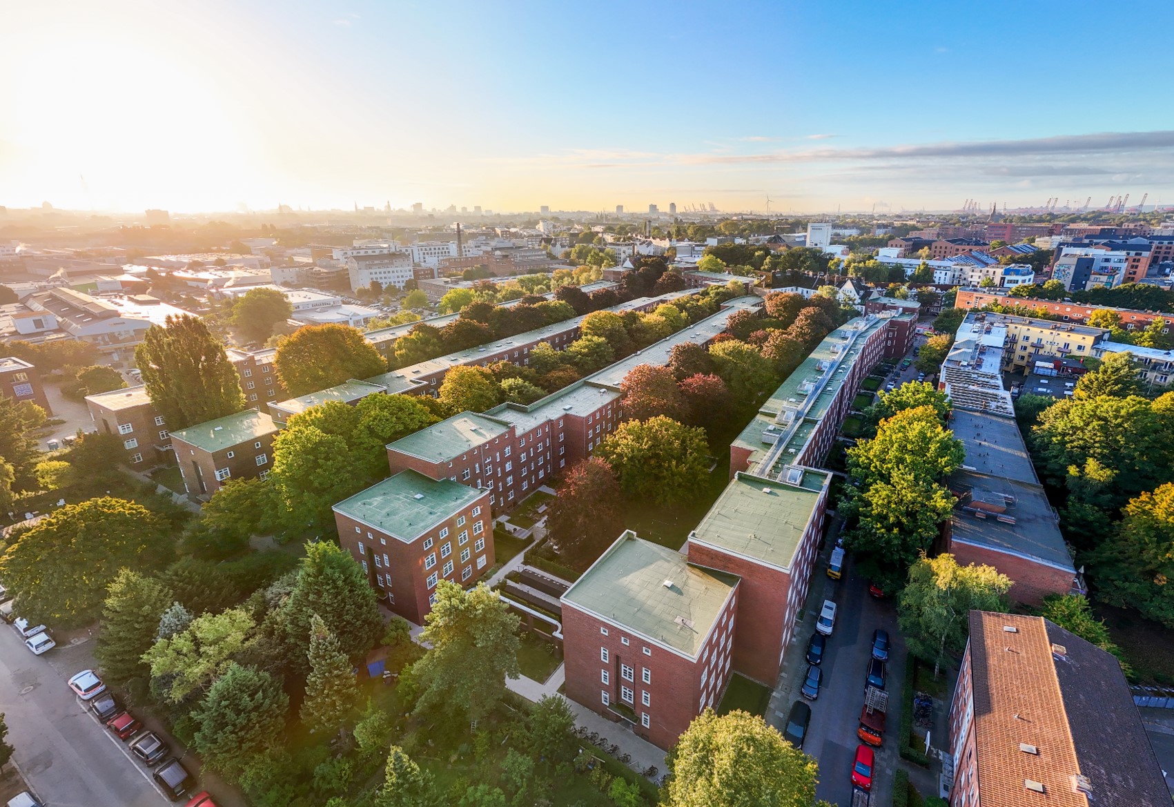 Luftaufnahme einer großen Wohnanlage mit mehreren viergeschossigen Backsteinbauten in U-Form, umgeben von Bäumen und Straßen; im Hintergrund Stadtansicht mit Industrie- und Hafenanlagen.