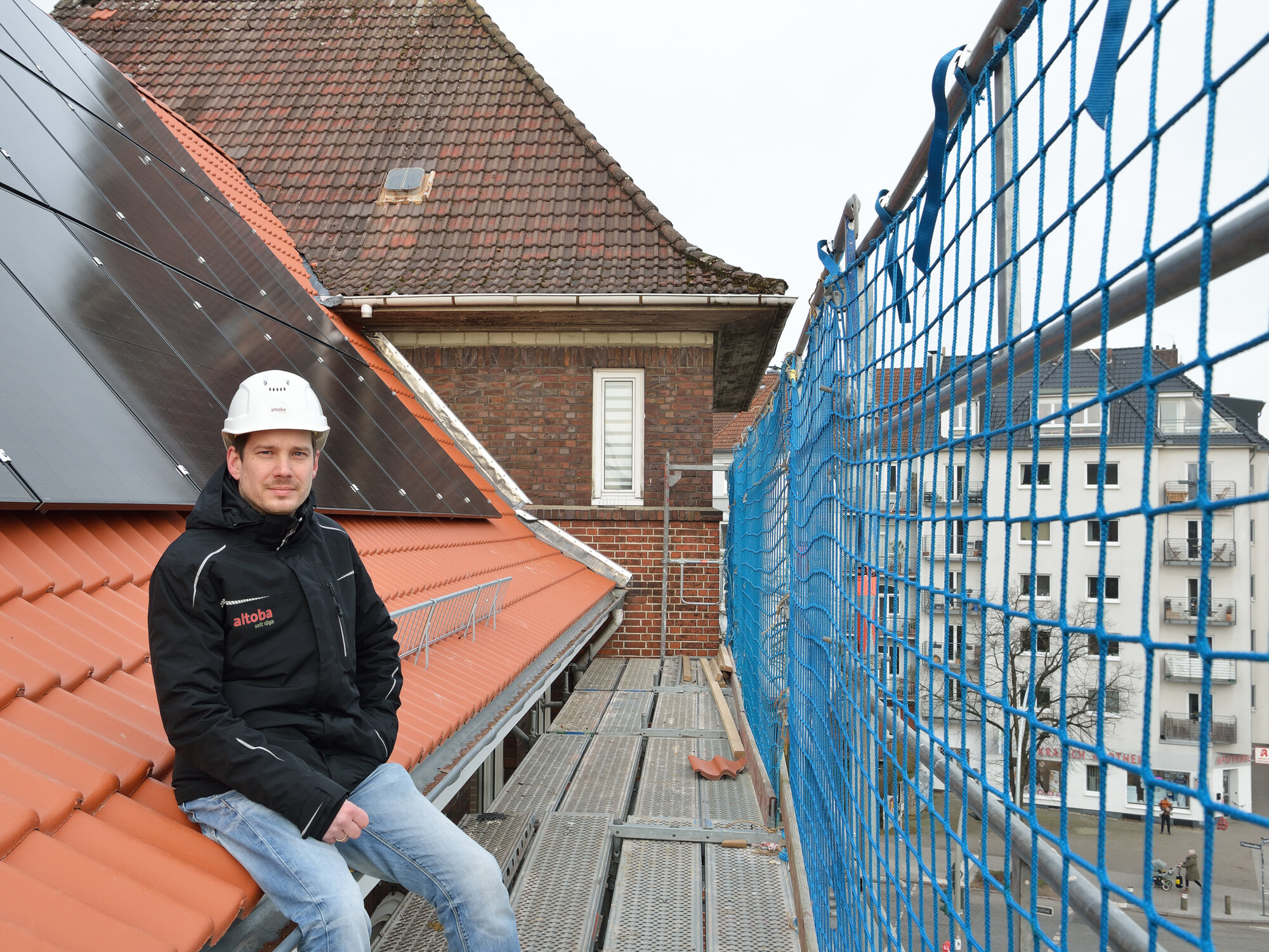Ein Mann mit Schutzhelm und altoba-Jacke sitzt auf einem Gerüst vor einem Ziegeldach mit installierten Solarmodulen; im Hintergrund ein weiteres Wohnhaus und eine Straße mit parkenden Autos und Passanten.