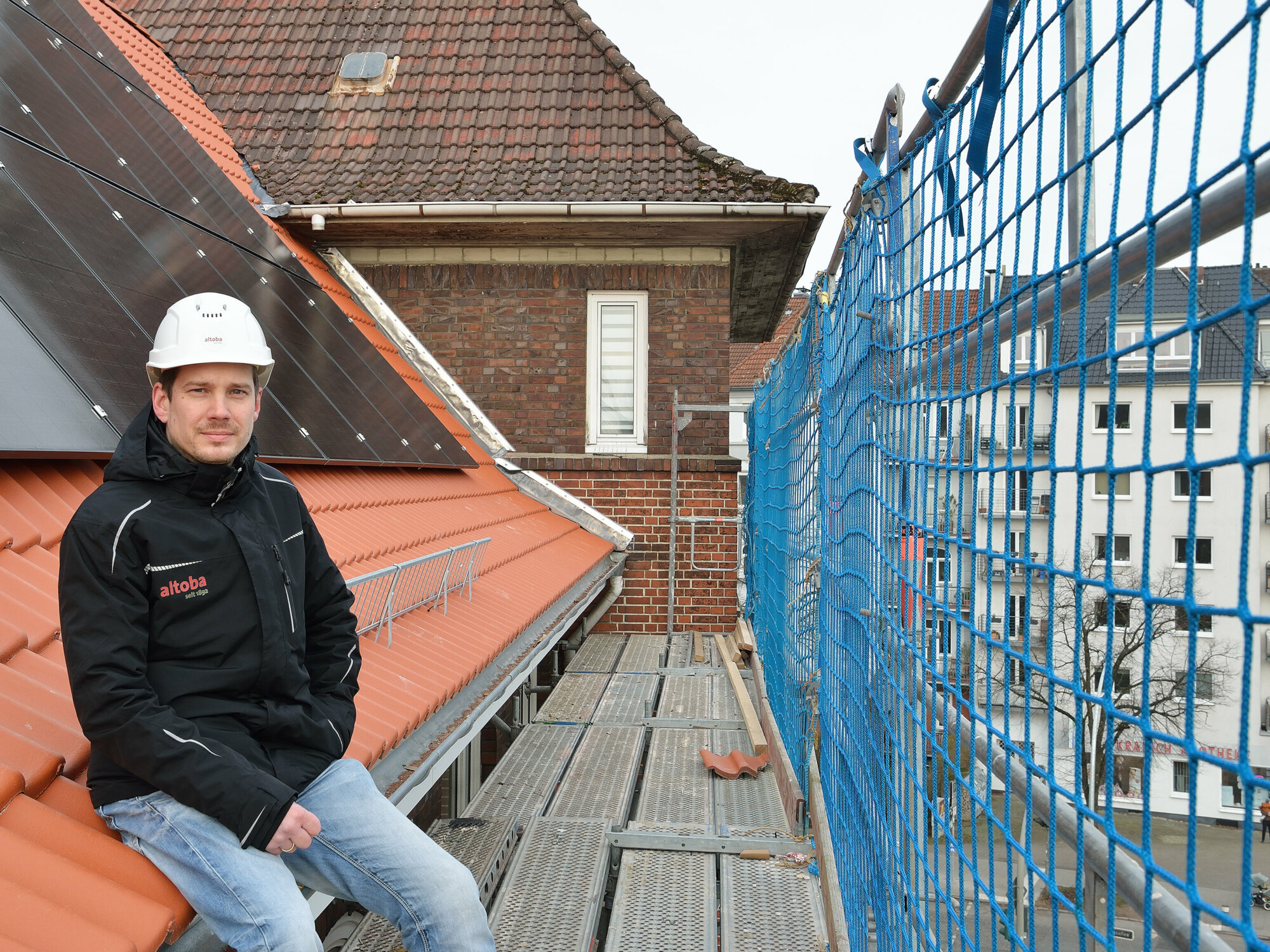 Ein Mann mit Schutzhelm und altoba-Jacke sitzt auf einem Gerüst vor einem Ziegeldach mit installierten Solarmodulen; im Hintergrund ein weiteres Wohnhaus und eine Straße mit parkenden Autos und Passanten.