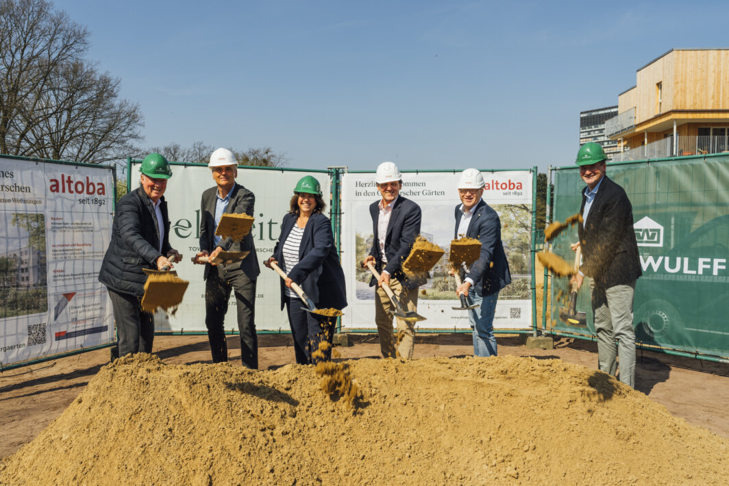 Sechs Personen mit Schutzhelmen werfen bei einem Spatenstich Sand in die Luft, vor Bauzaun mit altoba-Projektinformationen und Neubau im Hintergrund.