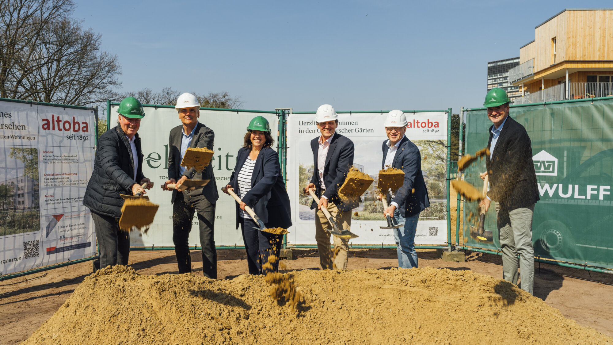 Sechs Personen mit Schutzhelmen werfen bei einem Spatenstich Sand in die Luft, vor Bauzaun mit altoba-Projektinformationen und Neubau im Hintergrund.