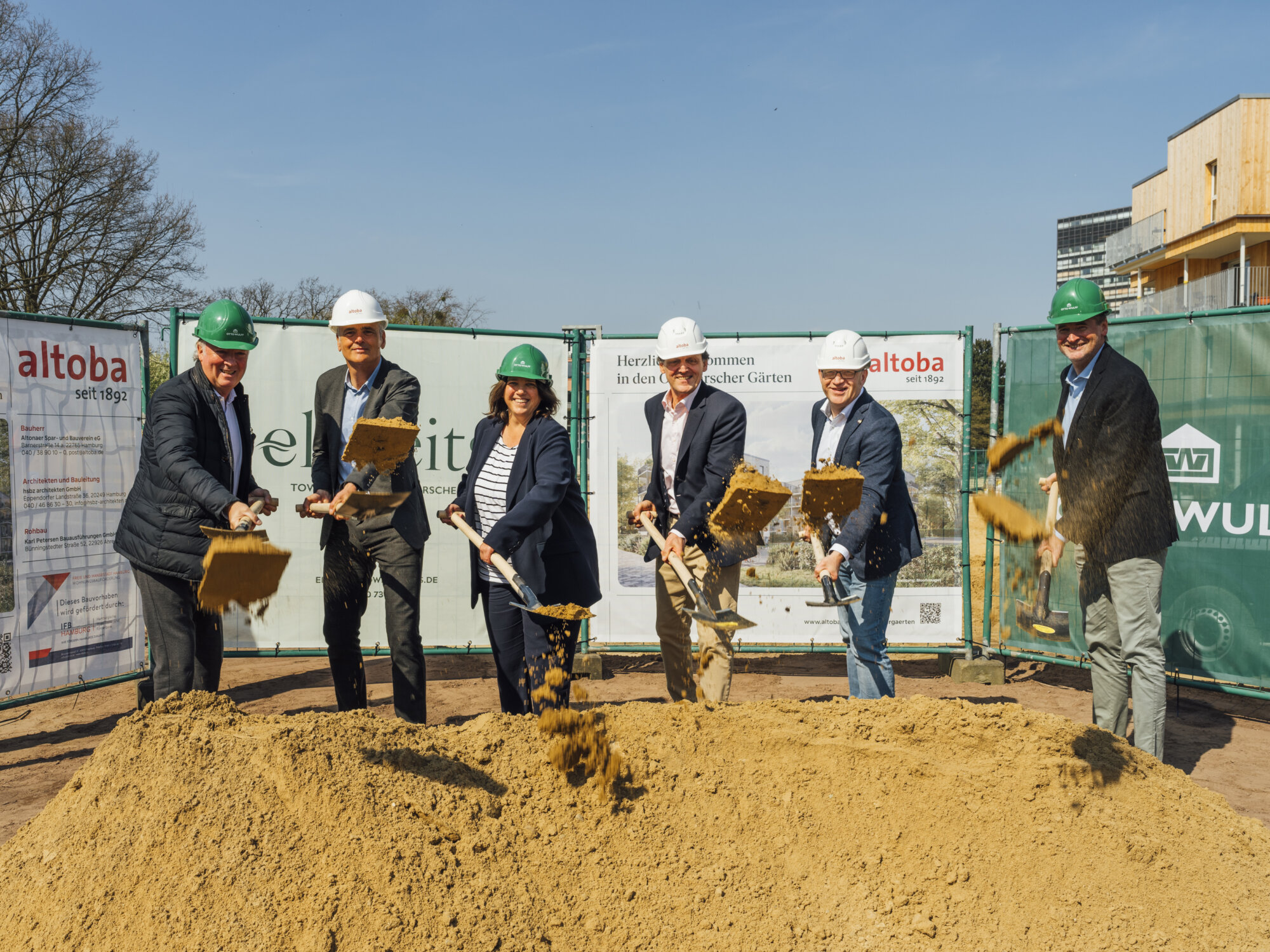 Sechs Personen mit Schutzhelmen werfen bei einem Spatenstich Sand in die Luft, vor Bauzaun mit altoba-Projektinformationen und Neubau im Hintergrund.