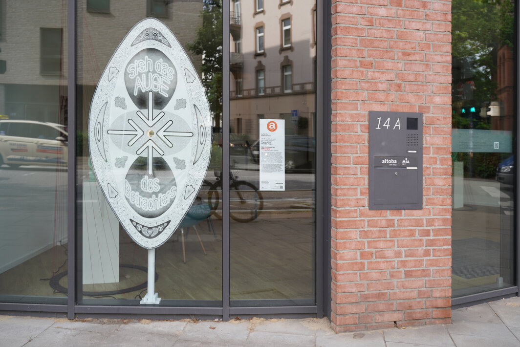 Außenansicht eines altoba-Gebäudes mit großer Schaufensterfront; vor dem Fenster steht ein Kunstobjekt mit der Aufschrift „sieh das AUGE des Betrachters“, daneben sind die Hausnummer 14A und eine Gegensprechanlage an einer Backsteinfassade sichtbar.
