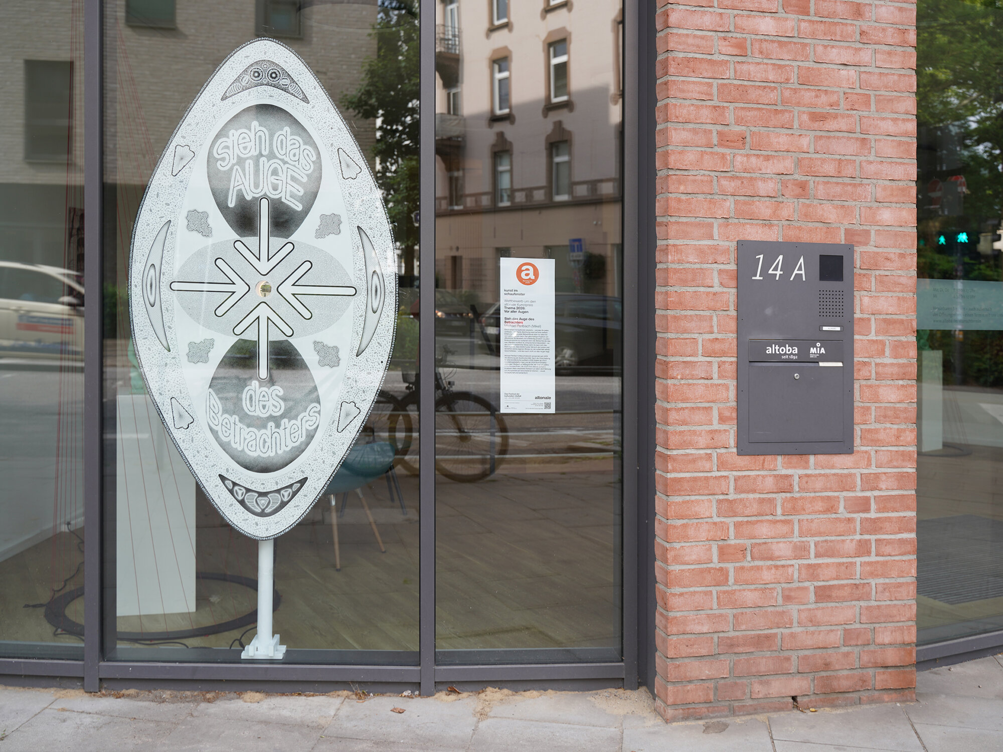 Außenansicht eines altoba-Gebäudes mit großer Schaufensterfront; vor dem Fenster steht ein Kunstobjekt mit der Aufschrift „sieh das AUGE des Betrachters“, daneben sind die Hausnummer 14A und eine Gegensprechanlage an einer Backsteinfassade sichtbar.